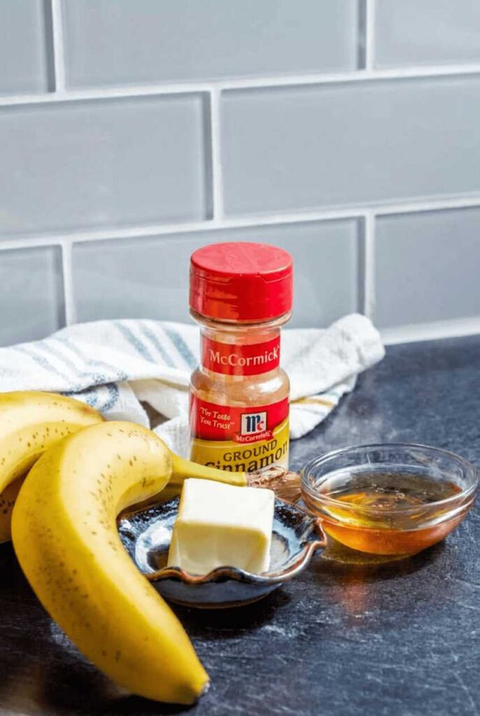 Fresh yellow bananas, a stick of butter, a jar of ground cinnamon, and a small bowl of maple syrup on a dark countertop.