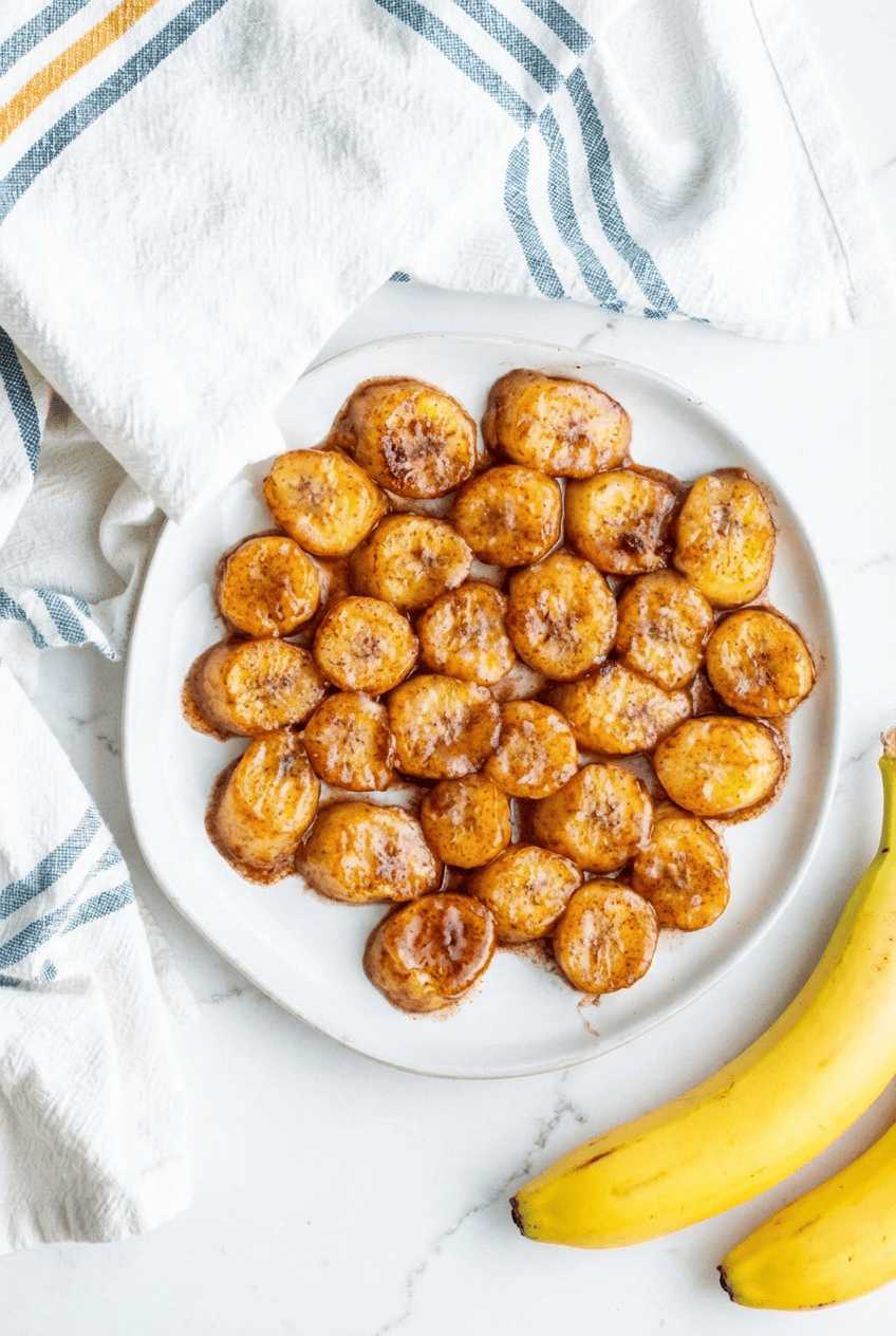 A white plate filled with circular pan-fried banana slices coated in a dark cinnamon glaze, served next to fresh bananas.
