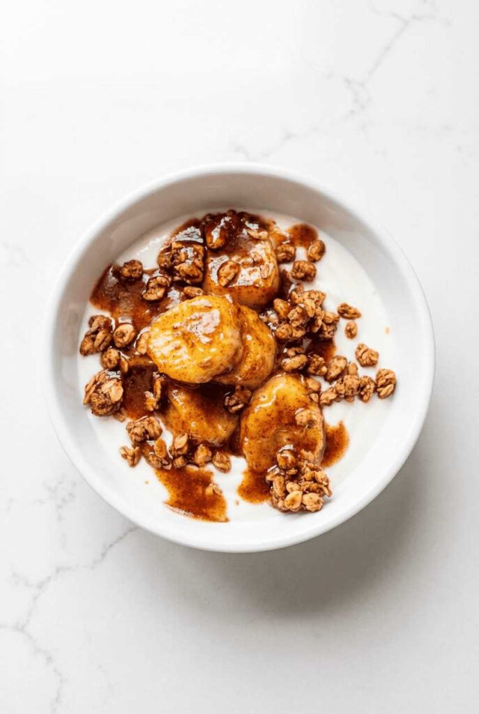 A white breakfast bowl containing Greek yogurt topped with granola and warm caramelized cinnamon bananas.