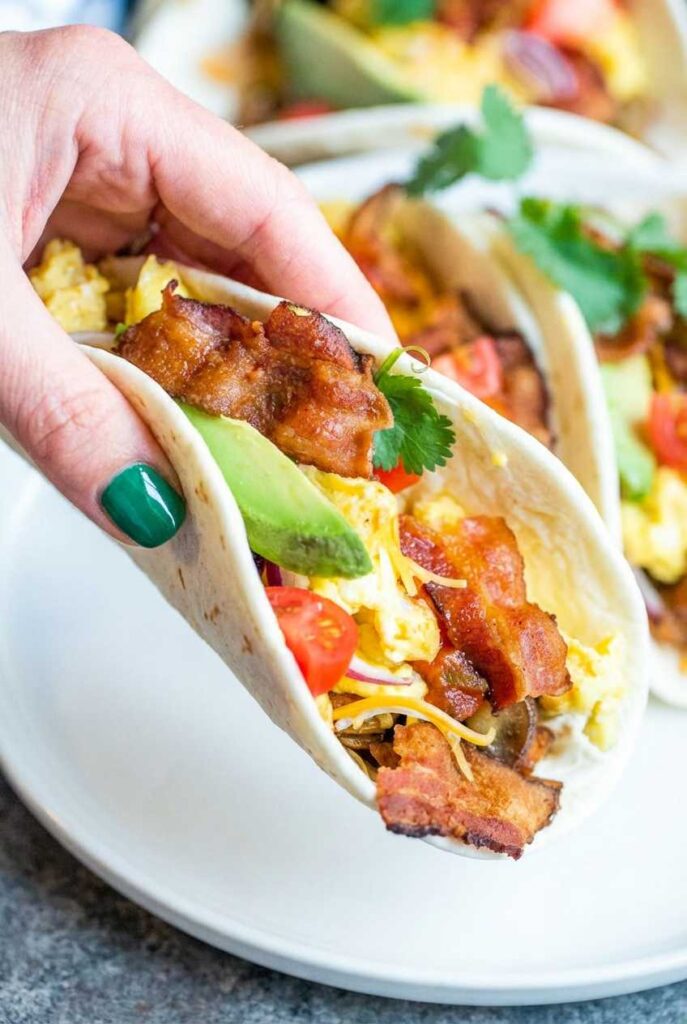 A hand holding a flour tortilla breakfast taco filled with scrambled eggs, crispy bacon, avocado slices, and fresh cilantro.