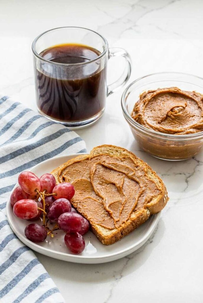 A slice of toast spread with smooth cookie butter served with red grapes and a mug of black coffee.