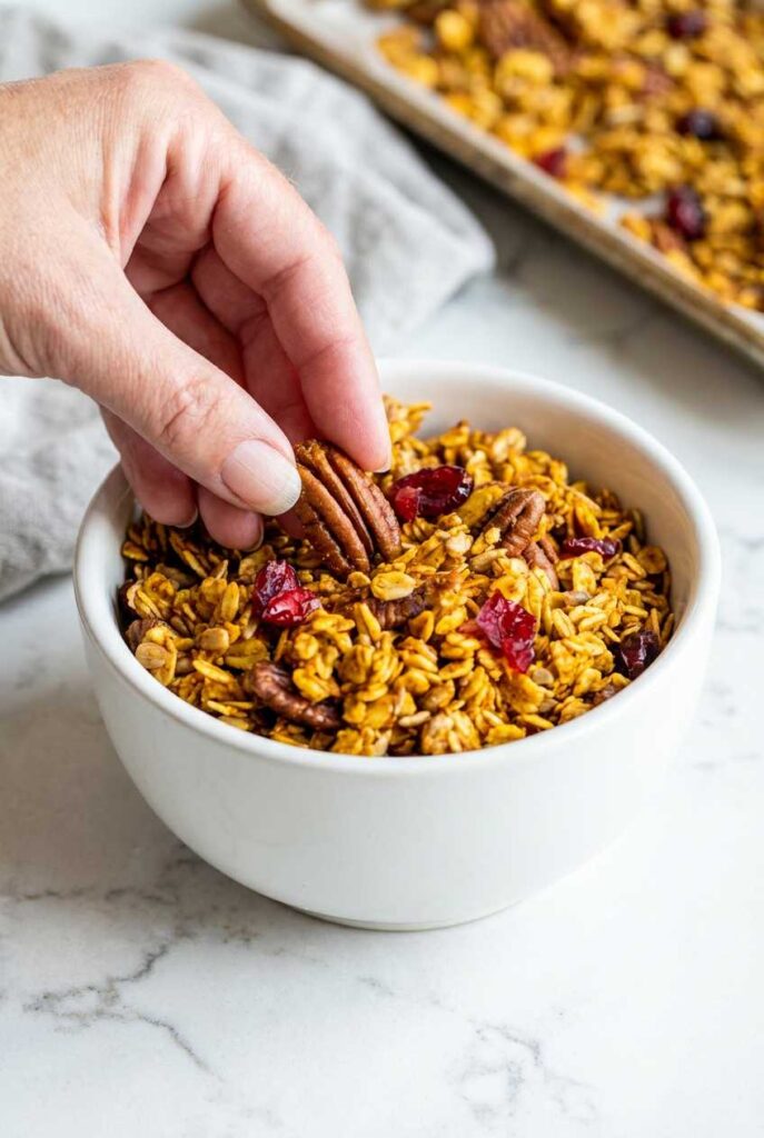 A hand placing a large pecan on top of a white bowl filled with golden-brown pumpkin spice granola and dried cranberries.
