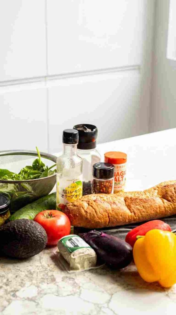 A collection of fresh ingredients on a marble countertop including a long baguette, a whole avocado, a ripe tomato, colorful bell peppers, an eggplant, and a bowl of fresh spinach.