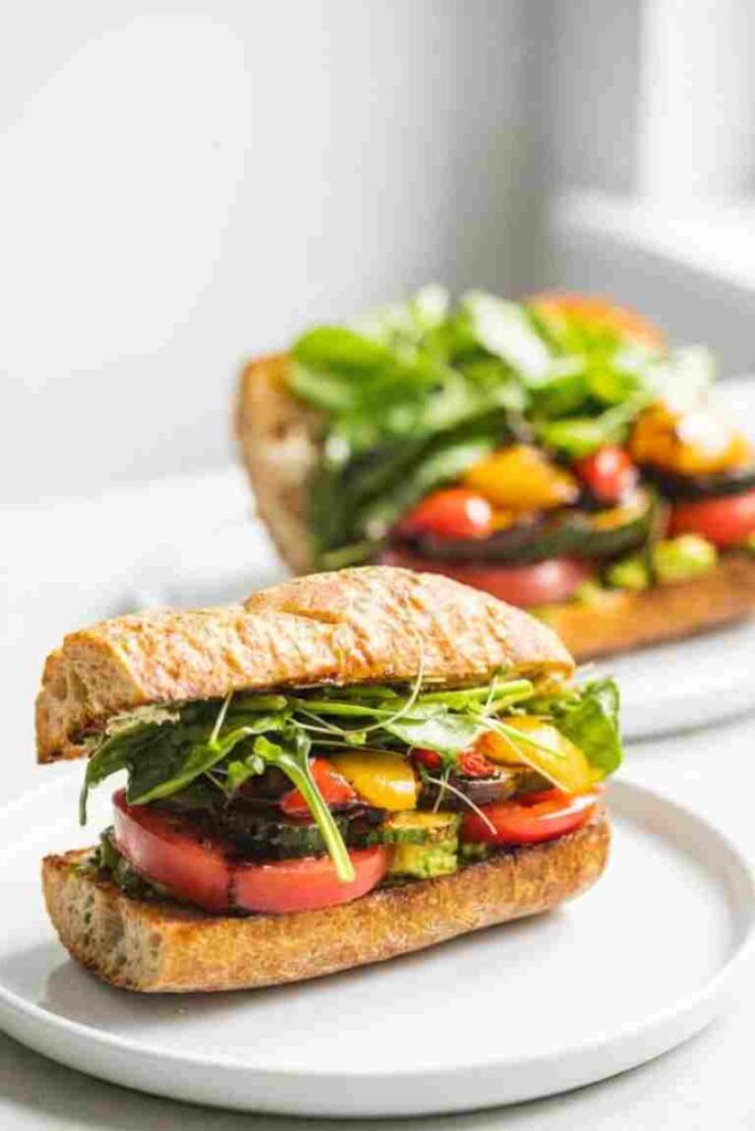 Two completed roasted vegetable sandwiches filled with peppers, zucchini, tomatoes, and greens on a white plate.