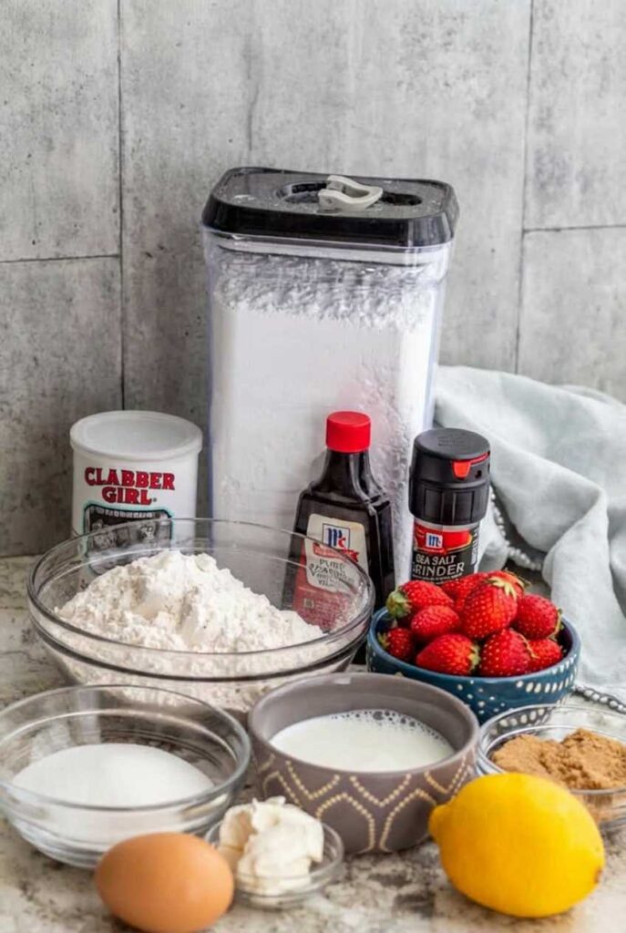 Baking ingredients including flour, sugar, eggs, milk, vanilla, a fresh lemon, and a bowl of ripe strawberries on a counter.