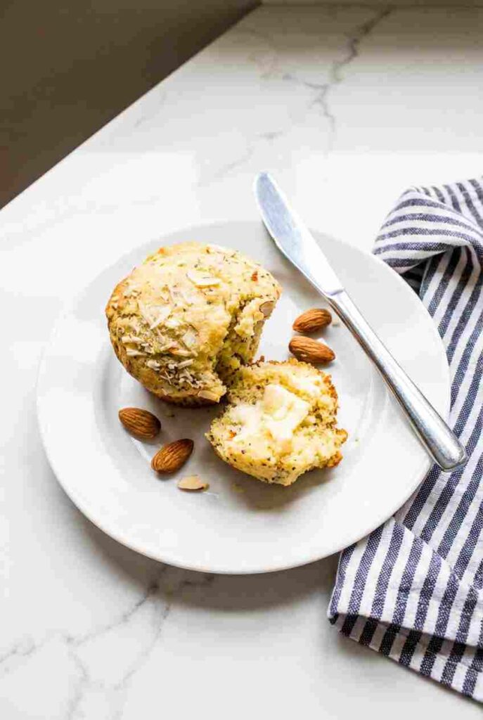 A single almond poppy seed muffin sliced open with a pat of melting butter, served on a white plate with whole almonds.