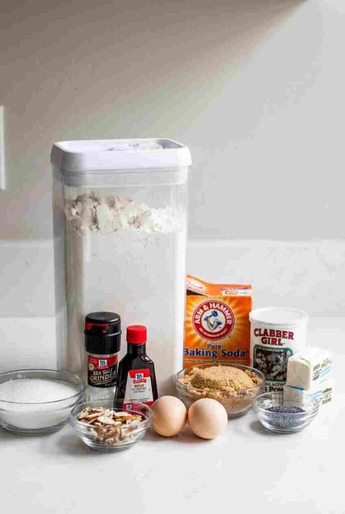 Baking staples including flour, sugar, eggs, butter, poppy seeds, vanilla, and almond slices arranged on a white counter.